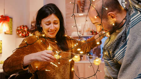 Couple laughing and getting tangled in string of christmas lights. Man and woman trying to untangle knot of garland with twinkle lights and illuminated bulbs. Holiday decorationsの写真素材
