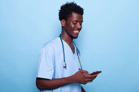 Man working as nurse looking at smartphone and smiling. Medical specialist wearing uniform and stethoscope holding modern phone with touch screen. Healthcare assistant using deviceの写真素材