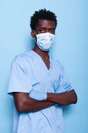 Nurse with crossed arms wearing face mask and gloves, looking at camera in studio. Man working as specialist standing over blue background, having protection against covid 19 pandemicの写真素材