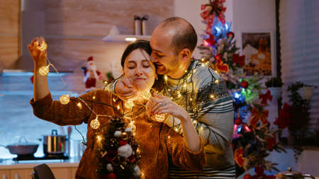 Festive couple knotted in string of christmas lights. Man and woman laughing while trying to untangle garland with twinkle lights and illuminated bulbs for holiday celebration.の写真素材