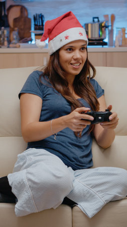 Woman playing video games on console with controller for christmas eve festivity. Adult with santa hat smiling and holding joystick for play on television. Cheerful person on holidayの写真素材