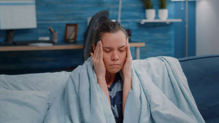 Close up of ill adult rubbing temples against headache. Sick woman with migraine sitting wrapped in...