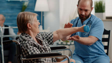 Patient with chronic disease doing physical exercises with dumbbells. Man nurse giving assistance to...