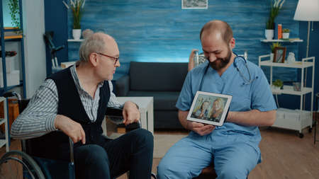 Retired man with disability talking to family on video call in nursing home while nurse holding tablet for assistance. Patient in wheelchair chatting on online remote conference with nieceの写真素材