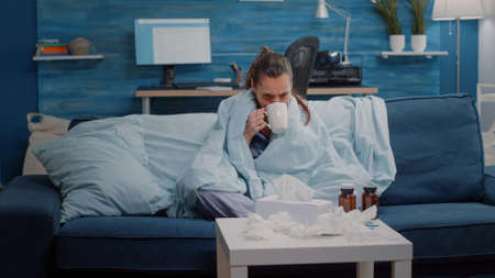 Adult shivering and feeling cold while using blanket and drinking tea for warmth. Woman with disease having chills and holding cup with hot beverage to cure coronavirus symptoms.の写真素材