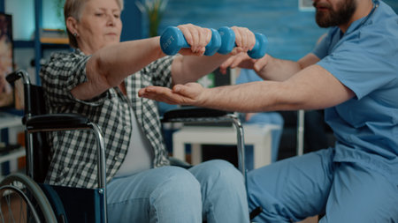 Close up of disabled woman using dumbbells for recovery therapy in nursing home. Medical assistant helping retired woman in wheelchair use objects for physical exercise and activityの写真素材