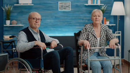 POV of senior couple with disability using video call to chat with family while sitting in nursing home. Man in wheelchair and woman with walk frame talking on online remote conferenceの写真素材
