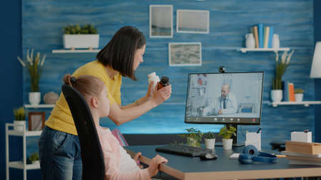 Mother using video call with doctor for healthcare treatment and advice. Parent holding bottle of pills asking medic on telemedicine about medication for telehealth and sick childの写真素材