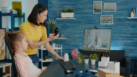 Mother asking doctor about bottle of pills on video call to cure ill child with fever. Parent and little girl doing online consultation with medic on video conference for telehealth and medicationの写真素材
