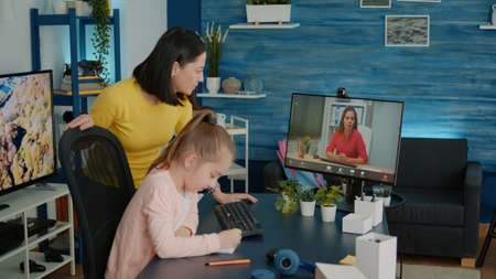 Child and mother using video call with teacher for online lesson and remote education. Young girl studying on video conference with professor for virtual course while parent giving helpの写真素材