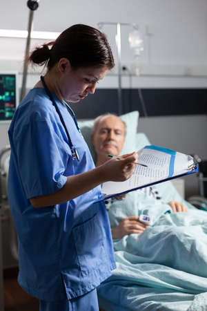 Medical nurse in scrubs taking notest on cliboard after consultation of sick unwell senior man. Patient with iv drip attached breathing with help from oxygen mask.の写真素材