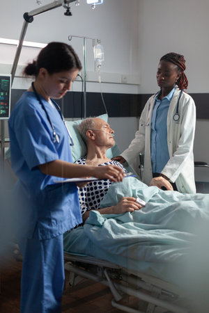 African doctor ward in hospital room discussing with sick senior man laying in bed breathing through oxygen tube with iv dirp bag attached, during consultation. Medical nurse taking notes on clipboard.の写真素材