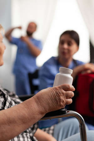 Disabled senior elderly patient holding vial of pills discussing disease therapy with support assistant worker. Woman nurse holding emergency red kit bag in hands. Social nusing services at homeの写真素材
