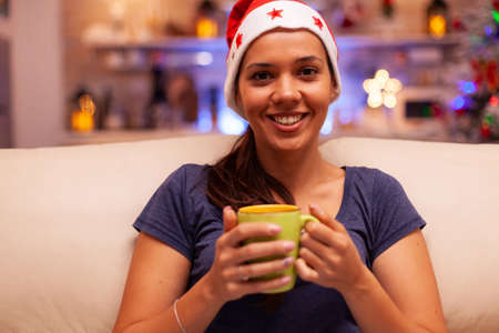 Portrait of woman wearing red santa hat holding cup of coffee in hands enjoying winter season relaxing on couch in xmas decorated kitchen. Adult celebrating christmas holidayの写真素材
