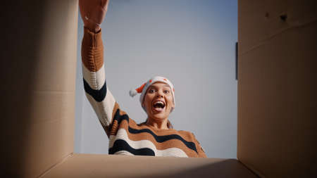 Caucasian woman with santa hat opening present box for surprise at home. Young festive adult smiling looking inside package. Person receiving christmas gift from friends and relativesの写真素材