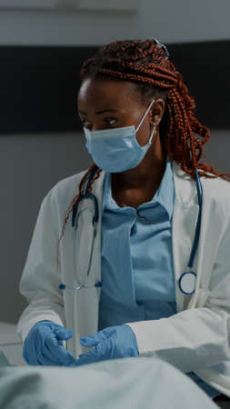 Recovering patient in hospital ward getting consultation from african american doctor with face mask and stethoscope. Elder man with nasal oxygen tube and oximeter sitting in bedの写真素材