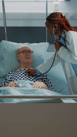 Multi ethnic personnel treats patient in hospital ward at medical clinic. Sick old man receiving modern treatment with nasal oxygen tube and IV drip bag from nurse and doctor at facilityの写真素材