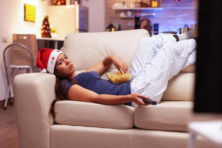Woman relaxing on couch eating snacks while watching xmas comedy movie on tv during christmastime in x-mas decorated kitchen. Adult person enjoying winter season celebrating christmas holidayの写真素材
