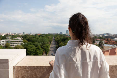 Woman tourist standing on tower terrace enjoying summer vacation looking at panoramic view of metropolitan city. Landscape with urban buildings seeing from rooftop observation point. Travel conceptの写真素材