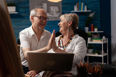Aged couple of artists highfive while using laptop for design inspiration in workshop space. Caucasian elder man and woman feeling cheerful and working on drawing for fine art projectの写真素材