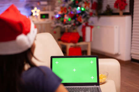 Woman sitting on couch in xmas decorated kitchen holding green screen mock up chroma key laptop computer with isolated display. Adult person enjoying christmas holiday celebrating winter seasonの写真素材