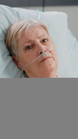 Portrait of senior patient with illness looking at camera while recovering from healthcare problem. Aged woman with nasal oxygen tube and oximeter for recovery in hospital ward bed.の写真素材