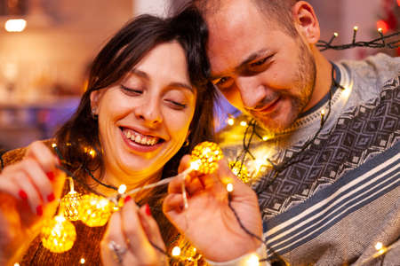 Amused couple stuck in christmas tree lights in xmas decorated kitchen celebrating christmas holiday. Happy family making funny while enjoying spending winter season together. New-year traditionの写真素材