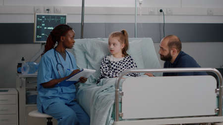 African american pediatrician woman nurse explaining healthcare treatment discussing sickness expertise during clinical examination in hospital ward. Sick child resting in bed after medicine surgeryの写真素材