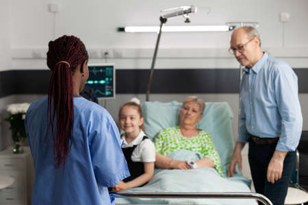 African american asisstant analyzing medical expertise explaining sickness treatment to elderly sick woman. Caring family checkup retired patient to support her during clincal therapy in hospital wardの写真素材
