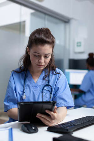 Practitioner nurse holding tablet computer analyzing sickness expertise working at healthcare treatment in hospital office. pediatrician asisstance checking pharmaceutical medication sitting at deskの写真素材