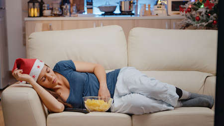 Sleepy woman with santa hat watching television on sofa. Festive adult falling asleep while looking...