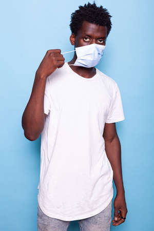 Portrait of person putting mask on face in studio. Casual man looking at camera while using hands to put face mask for protection against pandemic, standing over isolated backgroundの写真素材