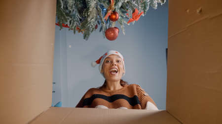 Young woman opening christmas present under tree being happy about surprise from friends and family. Caucasian person unpacking gift box while preparing for celebration festivityの写真素材