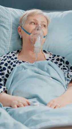 Elder woman receiving medical assistance from paramedics in hazmat suits using oximeter and oxygen tube to cure coronavirus symptoms in hospital ward. Quarantine area during pandemicの写真素材
