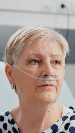 Close up of aged woman sitting in bed with heart rate monitor at clinic. Portrait of patient with illness using nasal oxygen tube for respiratory healthcare and recovery. Sick pensionerの写真素材