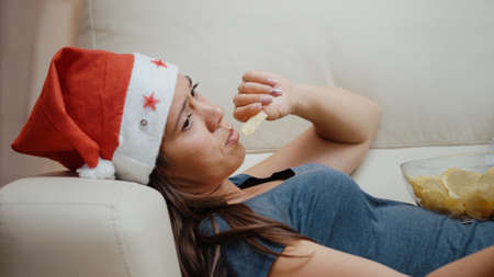 Bored woman wearing santa hat and eating chips from bowl while watching television and using TV remote control to switch channels on couch to celebrate christmas holiday. Person eatingの写真素材