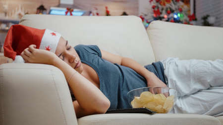 Close up of festive woman sleeping on sofa at television screen. Adult with santa hat resting on sofa while looking at movie on TV, feeling tired on christmas eve holiday. Sleepy personの写真素材