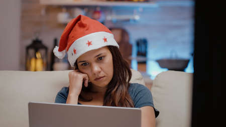 Close up of sleepy adult looking at laptop on christmas eve. Festive woman with santa hat falling asleep on couch while working with technology on device. Tired person on holidayの写真素材