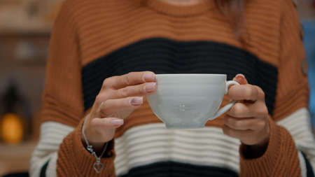 Close up of tea cup held by young woman at home in kitchen decorated with tree and ornaments. Adult preparing to drink from hot mug while waiting on christmas eve holiday dinner partyの写真素材
