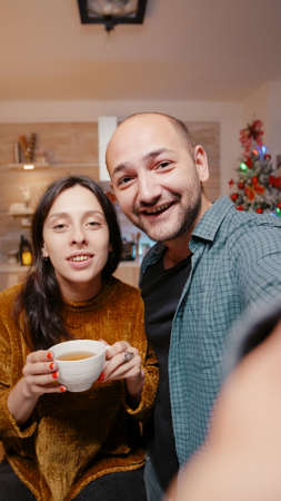 POV of festive couple talking on video call conference on christmas eve. Man and woman looking at camera smiling and chatting with relatives for holiday celebration at decorated home.の写真素材