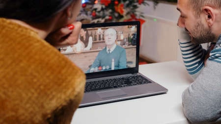 Festive couple talking to old man on online video call to celebrate christmas. People using conference for remote communication with elder person on laptop. Holiday festivity at homeの写真素材
