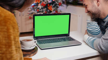Festive couple looking at horizontal green screen on christmas celebration. People watching chroma key with isolated background and mockup template on laptop for holiday festivity.の写真素材