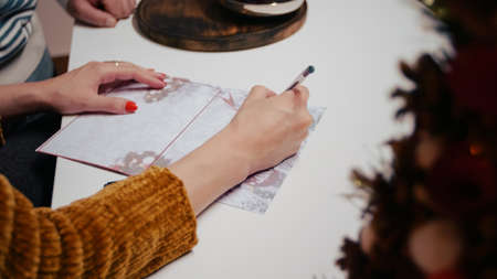 Close up of woman hand signing christmas gift card for holiday celebration. Adult writing on postcard paper for seasonal festivity, giving presents to relatives. Couple preparing for christmas.の写真素材