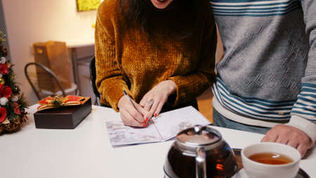 Close up of couple signing card for christmas festivity with family. Woman writing with pen on...