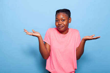 Portrait of confused african american teenager posing in pink t-shirt on a blue background in studio making uncertain facial expression. Young woman shrugging shoulders. I dont know conceptの写真素材