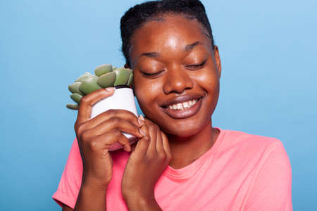 Closeup of of african american young woman enjoying gardening hobby holding flower pot in front of camera standing in studio with blue background. Portrait of teenager showing green flowerpotの写真素材