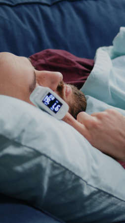 Close up of ill man sleeping on sofa with oximeter on finger. Adult using medical device with lower oxygen saturation measurement. Sick person with flu checking pulse pressure at homeの写真素材