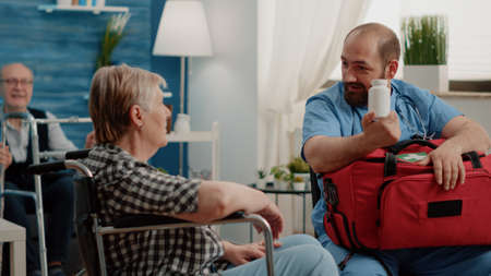 Old woman sitting in wheelchair receiving bottle of pils from man nurse for healthcare treatment and recovery. Specialist giving flask of medication to disabled patient in nursing homeの写真素材