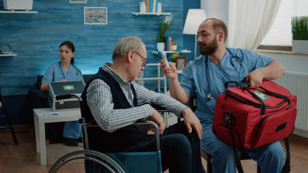 Medical assistant using thermometer for symptoms checkup with disabled man in nursing home. Nurse measuring temperature of senior patient in wheelchair for healthcare examination.の写真素材