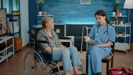 Nurse giving assistance to retired woman in nursing home. Caretaker at facility reading book for old disabled patient in wheelchair, medical assistant comforting person with chronic diseaseの写真素材